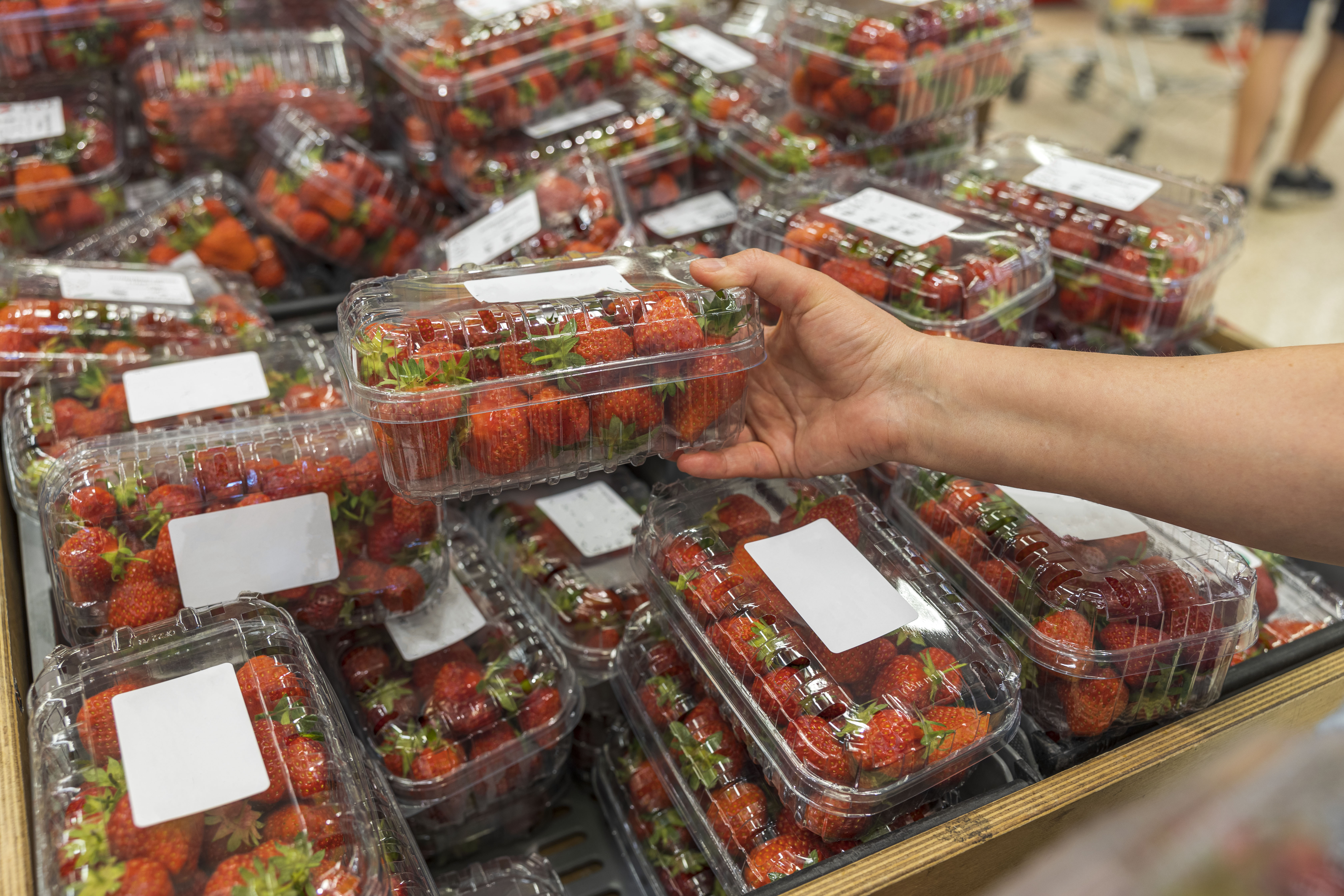 A hand is grabbing a punnet of strawberries from a display in a supermarket. A trolley can be seen in the distance.